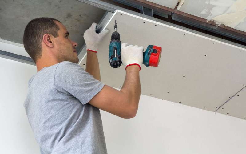 Construction worker assemble a suspended ceiling with drywall and fixing the drywall to the ceiling metal frame with screwdriver.